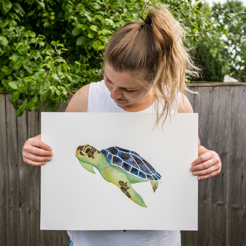 Jem standing in her garden, holding her original watercolour painting of a sea turtle